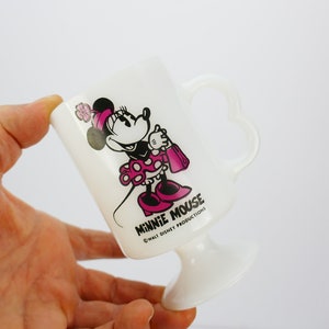 May include: White Minnie Mouse pedestal mug with a heart-shaped handle. The mug features a Minnie Mouse graphic in pink and black, holding a pink purse. The text "MINNIE MOUSE" and "&copy; WALT DISNEY PRODUCTIONS" are printed on the mug.