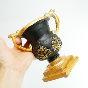 Peut inclure: Une urne décorative noire et dorée avec un motif de feuilles. L'urne a une base large et un col étroit avec deux poignées.