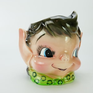 Könnte beinhalten: Eine Keramikfigur eines Elfen mit grün-weiß gepunkteter Kleidung. Der Elf hat braunes Haar, blaue Augen und ein verschmitztes Lächeln.