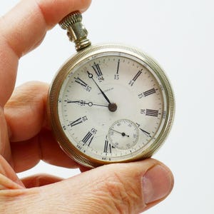 May include: A silver-coloured Waltham pocket watch with a white face, black Roman numerals, and a small seconds dial. The watch has a crown at the top. The word "Waltham" is printed on the watch face.