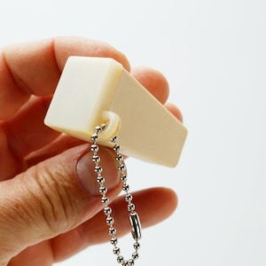 Puede incluir: Un silbato rectangular blanquecino con una cadena de bolas plateada. El silbato tiene una forma rectangular redondeada y un pequeño lazo para la cadena. La cadena está hecha de pequeñas bolas metálicas plateadas.