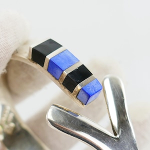 Peut inclure: Bague en argent ornée d'une incrustation rectangulaire de pierres précieuses noires et bleues alternées. Les pierres sont disposées en motif géométrique, contrastant avec le métal argenté. Fond blanc.
