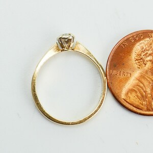 K&ouml;nnte beinhalten: Ein Goldring mit einem einzigen runden Diamanten, der in der Mitte eingesetzt ist. Der Ring wird neben einem US-Penny zur Gr&ouml;&szlig;envergleich gezeigt.