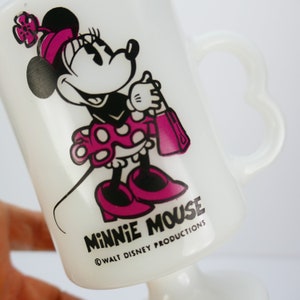 May include: White glass Minnie Mouse mug featuring a cartoon Minnie Mouse in a pink polka dot dress, shoes, and bow. The mug has a heart-shaped handle and the words "MINNIE MOUSE" and "&copy; WALT DISNEY PRODUCTIONS" printed on the front.