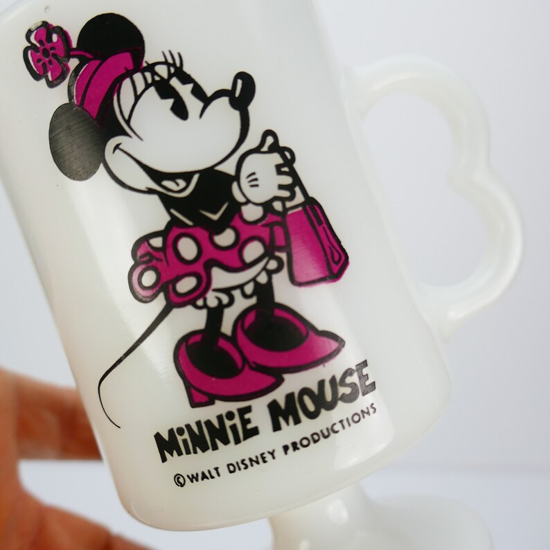May include: White glass Minnie Mouse mug featuring a cartoon Minnie Mouse in a pink polka dot dress, shoes, and bow. The mug has a heart-shaped handle and the words "MINNIE MOUSE" and "&copy; WALT DISNEY PRODUCTIONS" printed on the front.