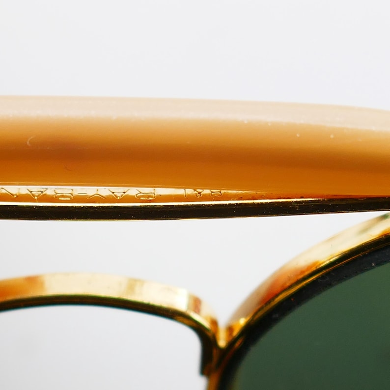 May include: Close-up of sunglasses with a gold frame and green lenses. The top of the frame is a light brown color. The text "RAY-BAN" is visible on the frame.