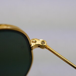 May include: Close-up of gold-rimmed sunglasses with dark green lenses. The hinge and arm of the glasses are visible, showcasing the gold metal's shine. The lenses have a slight curve, and the overall design suggests a classic style.