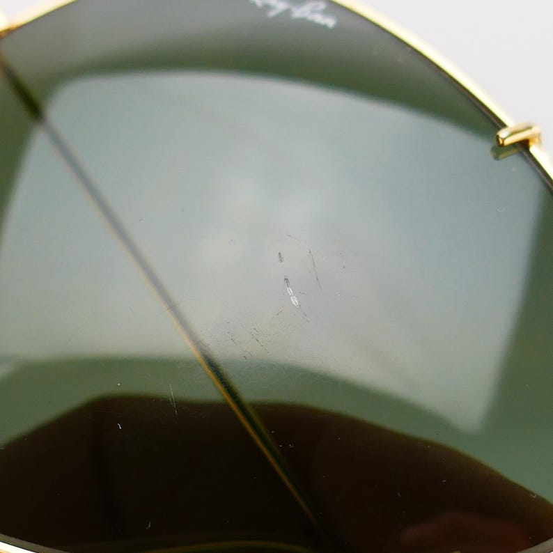 May include: Close-up of Ray-Ban sunglasses with a gold frame and green lenses. The lenses have a slight gradient and show minor scratches. The Ray-Ban logo is visible on the top left lens.