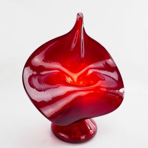 Puede incluir: Una vibrante escultura de vidrio rojo con una forma única y orgánica. La pieza presenta una parte superior acampanada y una base redondeada, creando un diseño dinámico y llamativo. La superficie brillante refleja la luz, realzando su atractivo visual. La escultura mide aproximadamente 25 cm de altura.