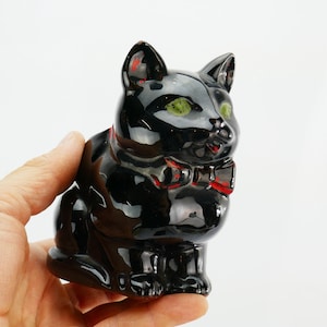 May include: A glossy black ceramic cat figurine with green eyes and a red bow tie. The cat is in a seated position, with its ears perked up. The figurine has a vintage or antique appearance.
