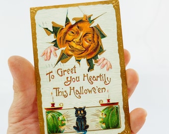 Könnte beinhalten: Vintage Halloween-Postkarte mit einem lächelnden Kürbisgesicht, das durch einen hölzernen Hintergrund bricht. Die Karte zeigt eine schwarze Katze, grüne Jack-o'-Laternen und den Text "To Greet You Heartily This Halloween."