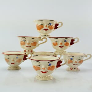 May include: A collection of cream-colored teacups, each adorned with a whimsical face design. The faces feature blue eyes, red lips, and orange cheeks. The teacups have a red trim and a decorative handle. The teacups are stacked on top of each other.