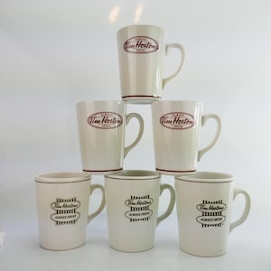 May include: A stack of seven off-white ceramic mugs. The mugs have a handle and a dark red rim. The mugs have the Tim Hortons logo and the words "ALWAYS FRESH" printed on them.