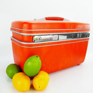 Puede incluir: Una maleta de viaje naranja vintage con ribetes plateados y un asa. La maleta tiene una superficie texturizada y un cierre plateado. Dos limas y dos limones están delante de la maleta.