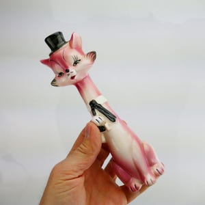 May include: A pink and white ceramic cat figurine wearing a black top hat and a black bow tie. The cat has a long neck and is holding a small object in its paws. The figurine has black and white accents.