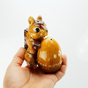 Könnte beinhalten: Ein Salz- und Pfefferstreuer-Set aus Keramik in Form eines Eichhörnchens. Das Eichhörnchen ist braun und hellbraun und hält einen braunen, eichelförmigen Shaker. Das Set wird in einer Hand vor weißem Hintergrund gehalten.