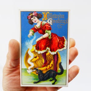 Peut inclure: Carte postale vintage d'Halloween représentant une femme en robe rouge sur une citrouille sculptée. Un chat noir boit dans une tasse. La carte postale porte les mots "Halloween Greetings" dans une police décorative sur fond bleu.