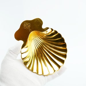 Peut inclure: Un plat décoratif doré en forme de coquillage. Le plat présente une croix sur le dessus et un motif nervuré à l'intérieur. Le plat est tenu par une main gantée sur fond blanc.