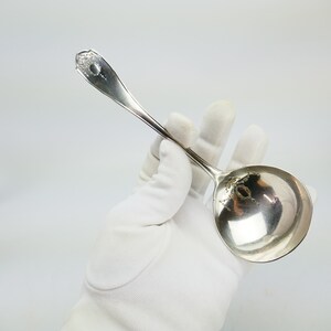 Puede incluir: Una cuchara de servir de plata con un mango adornado y un cuenco redondo. La cuchara se sostiene en una mano enguantada de blanco.