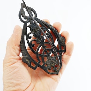 May include: Ornate, dark metal hair barrette with a decorative floral and scroll design. The barrette features a curved, openwork frame with a central pin for securing hair. The metal has an aged, antique appearance.