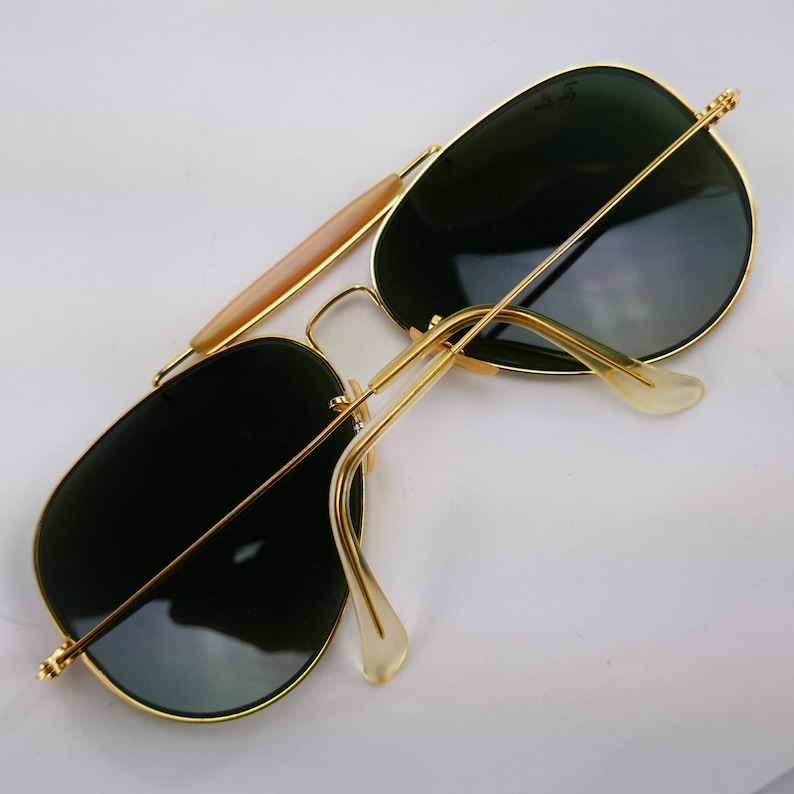 May include: Gold-framed sunglasses with dark green lenses. The sunglasses have a double bridge and a light-colored bar across the top of the frame. The temples are thin and gold, with clear ear pieces.
