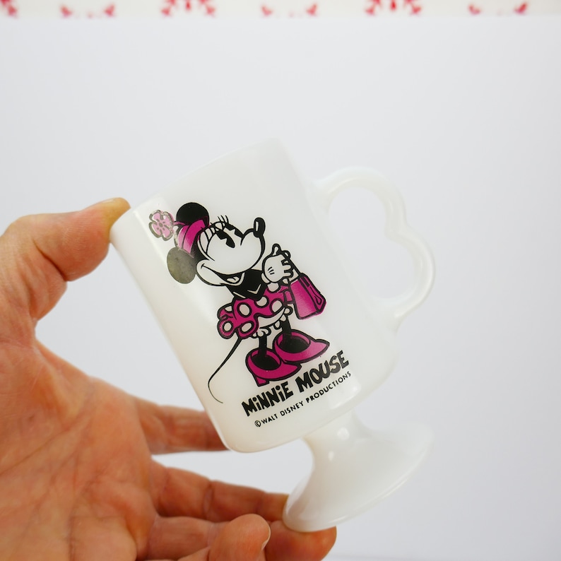 May include: White Minnie Mouse pedestal mug with a heart-shaped handle. The mug features a Minnie Mouse graphic in pink and black, holding a pink purse. The text "MINNIE MOUSE" and "&copy; WALT DISNEY PRODUCTIONS" are printed on the mug.