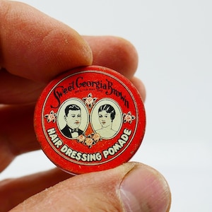 Puede incluir: Una lata roja vintage de Sweet Georgia Brown Hair Dressing Pomade. La lata circular presenta dos retratos ovalados y el nombre del producto en una fuente decorativa. El texto "Hair Dressing Pomade" también está impreso en la lata.