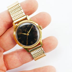 May include: A vintage Elgin wristwatch with a gold-tone case and a black dial. The watch features gold hands and hour markers, with the brand name "ELGIN" displayed. The watch band is a gold-tone, expandable style.