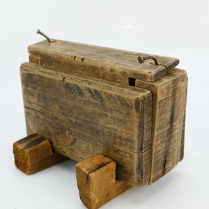 May include: A weathered wooden box with a hinged lid, made from aged planks. The box is supported by two wooden blocks, and secured with metal hardware. The wood has a natural, aged appearance, with visible grain and knots.