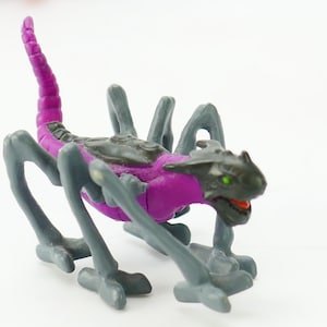 Pode incluir: Uma criatura de brinquedo de plástico roxo e preto com várias pernas e uma cauda longa. A criatura tem uma cabeça de dragão com um olho verde.