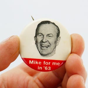 May include: A vintage campaign badge featuring a smiling man's portrait. The badge has a cream background with a red stripe at the bottom, reading "Mike for me in '63".