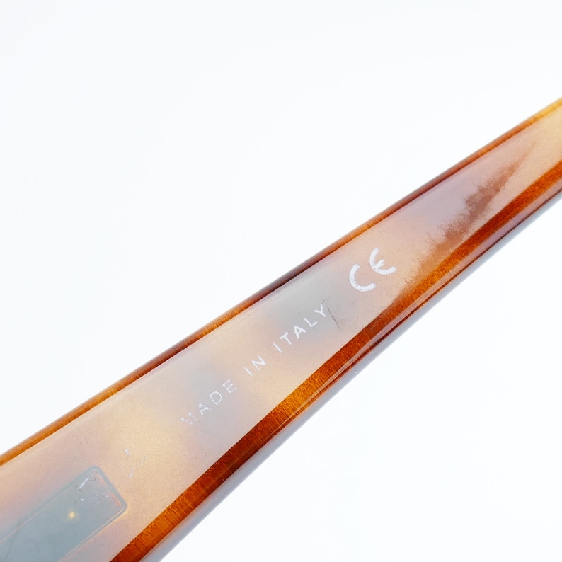 Peut inclure: Gros plan sur la branche en &eacute;caille brune d'une paire de lunettes. La branche est marqu&eacute;e du texte "MADE IN ITALY" et du marquage CE.