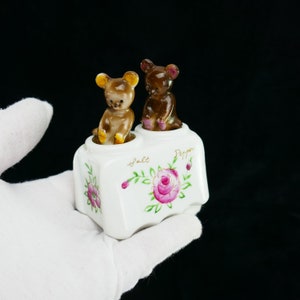 Puede incluir: Juego de salero y pimentero de cerámica blanca con diseño floral y dos figuritas de ositos de peluche marrones en la parte superior. Los shakers están etiquetados como "Sal" y "Pimienta".