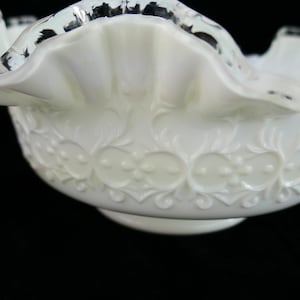 Puede incluir: Un cuenco blanco con borde ondulado y diseño floral en relieve. El cuenco tiene un borde festoneado con detalles negros. El patrón detallado incluye elementos circulares y en forma de hoja, creando una estética vintage. El cuenco se apoya en una pequeña base circular.