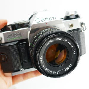 Könnte beinhalten: Eine silberne und schwarze Canon AE-1 Program Kamera mit einem 50mm 1:1.8 Objektiv. Das Canon-Logo ist grün und das Objektiv trägt die Aufschrift "CANON LENS FD 50mm 1:1.8 MADE IN JAPAN". Die Kamera wird in einer Hand gehalten.