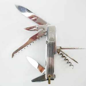 May include: A multi-tool knife with various stainless steel blades and tools, including a corkscrew, saw, and knife blades. The knife has the word "THIERS" printed on the side.