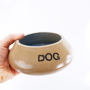 May include: Tan ceramic dog bowl with a blue interior. The word "DOG" is printed in black on the side of the bowl. The bowl is held by a hand against a white background.
