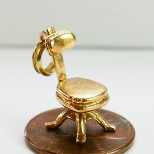 May include: A gold-toned charm in the shape of an office chair. The chair has a rounded back, a square seat, and four legs. The charm is about the size of a US penny.