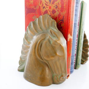 Op de afbeelding: Bronzen paardenkop boekensteunen. De boekensteunen staan naast een rij vintage boeken met rode, blauwe en groene covers. De paardenkop heeft gedetailleerde kenmerken en een verweerde uitstraling, wat charme toevoegt.