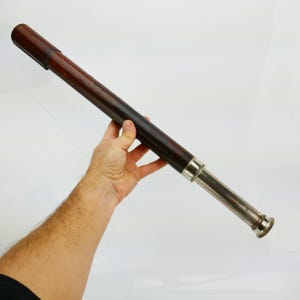 May include: A vintage telescope with a dark brown leather-wrapped body and a silver-colored metal eyepiece. The telescope is held at an angle against a white background. The leather has a rich, aged appearance.