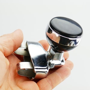 May include: A chrome and black steering wheel spinner knob. The knob has a shiny black top and a chrome base. The clamp is also chrome. The object is held in a hand against a white background.