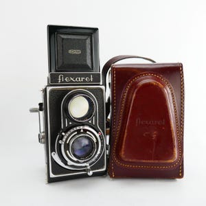 May include: A vintage Flexaret twin-lens reflex camera with a black body and silver accents. The camera has a top viewfinder and a lens with various settings. A brown leather case with the Flexaret logo is next to the camera.