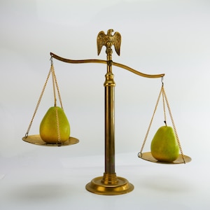 May include: A vintage brass balance scale with an eagle emblem on top, holding two green pears on its trays. The scale is set against a white background, highlighting the contrast between the fruit and the antique metal.