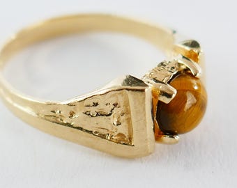 May include: A gold-colored ring with a textured band and a round, brown tiger's eye stone. The ring's setting has a textured design, and the stone is securely held in place by prongs.