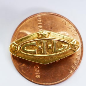 May include: A gold-colored metal pin with the letters "C.J.L." in a diamond-shaped design. The pin has two stars on either side of the letters.