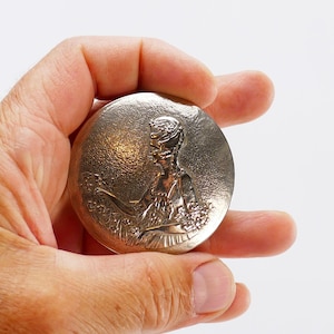 Könnte beinhalten: Eine silberfarbene, runde Medaille, die in einer Hand gehalten wird. Die Medaille zeigt ein detailliertes Relief einer Frau in klassischer Kleidung, die Blumen hält. Die Oberfläche hat ein strukturiertes, gehämmertes Aussehen, und das Gesamtdesign ist verziert.