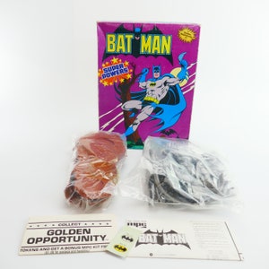 Puede incluir: Una caja de kit de modelo vintage de Batman con un fondo morado y una imagen de Batman con una capa azul. La caja también incluye un kit de modelo de plástico rojo y marrón, una bolsa de piezas de plástico negro y un pequeño folleto de instrucciones con las palabras "Collect Golden Opportunity" y "MPC BAT MAN" impresas en él.