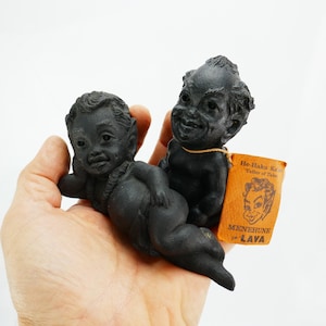 May include: Two black Menehune figurines, one reclining and one upright, held in a hand. The figurines have detailed facial features and are made of a dark material. A small tag reads "He-Haku Ka Teller of Tales" and "Menehune in LAVA."