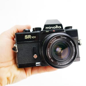 May include: A black Minolta SR 101 camera with a Vivitar wide-angle lens. The lens has a 49mm diameter and a 28mm focal length. The camera body has the text "SR 101" and "minolta CLC". The camera is held in a hand against a white background.