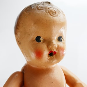Puede incluir: Una muñeca bebé vintage con la cara pintada. La muñeca tiene ojos negros, mejillas rojas y labios rojos. La piel de la muñeca es de color melocotón claro, y el pelo está moldeado en un remolino en la parte superior de la cabeza. Los brazos de la muñeca son visibles.